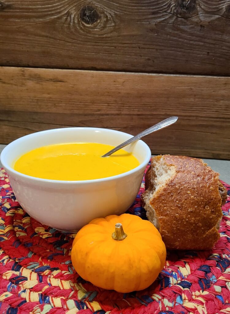 Pumpkin soup in a white bowl with bread and a small pumpkin would make a great additions for easy Yule dinner ideas and recipes for a magical feast. 
