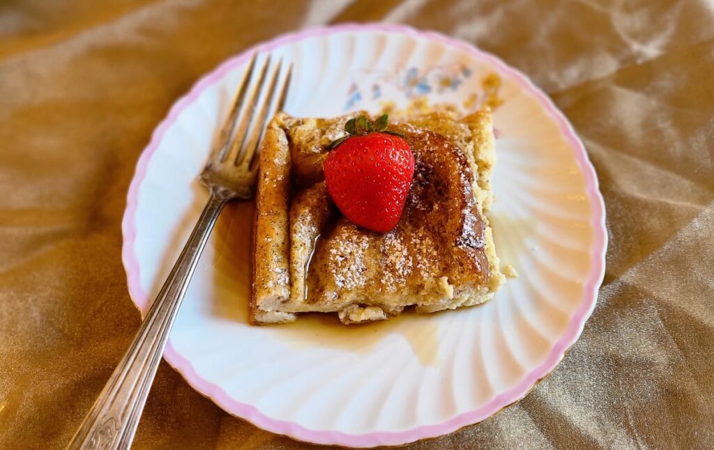 Pretty plate with a piece of cardamom French toast with a strawberry on top with a silver fork.