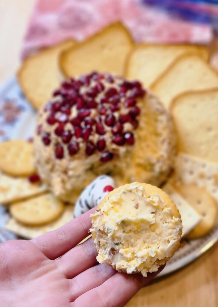 Hand with a cracker and cheese in front of an easy low carb cheese ball with mascarpone and gouda