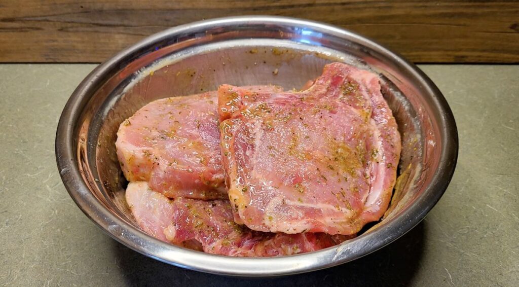 Raw pork chops taking a bath in a marinade in a metal bowl. 