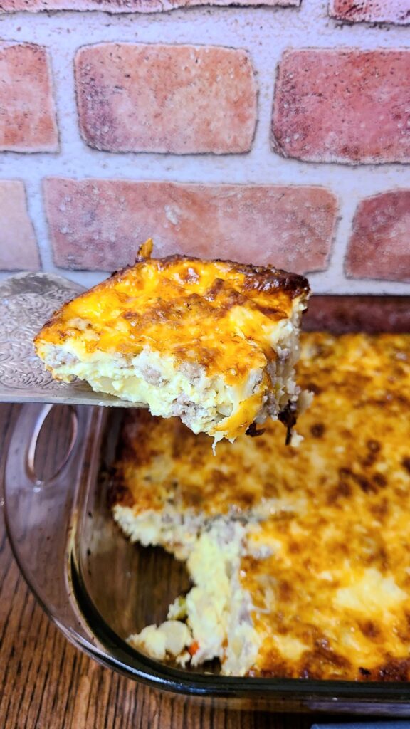 Piece of Easy overnight sausage, egg, & potato breakfast casserole on a silver spatula with a brick background. 