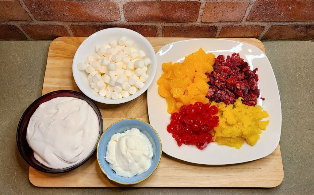 Ingredients to make  how to make ambrosia fruit salad with whipped cream laid out on a wood cutting board.
