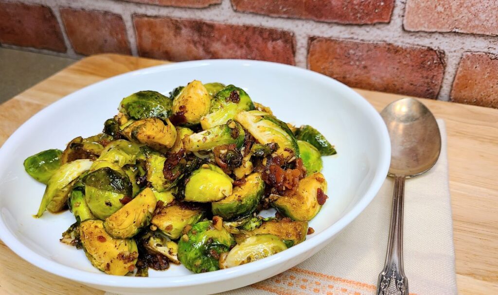 easy sauteed maple & balsamic glazed bacon brussels sprouts in a white bowl with a spoon and cloth napkin.